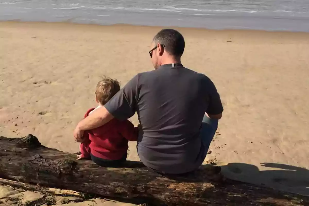 Un adulto y un niño sentados en un tronco en la playa mirando hacia el mar. Un adulto y un niño sentados en un tronco en la playa mirando hacia el mar.