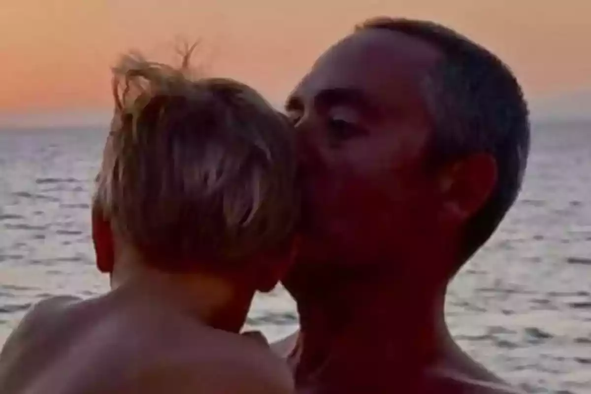Un adulto sostiene y besa a un niño pequeño frente al mar durante el atardecer. Un adulto sostiene y besa a un niño pequeño frente al mar durante el atardecer.