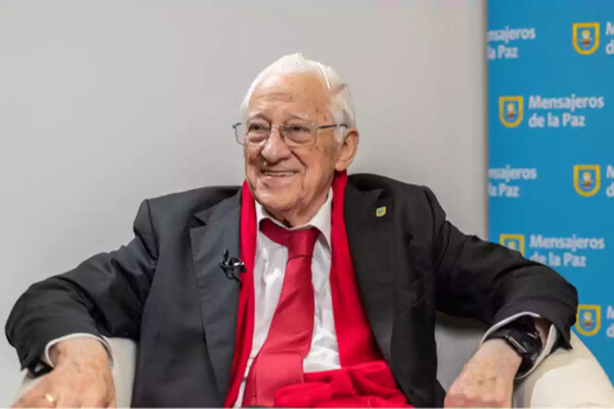 Un hombre mayor con traje y corbata roja sonríe mientras está sentado en una silla con un fondo que tiene el logo de "Mensajeros de la Paz".