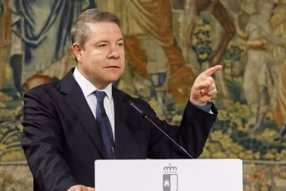 Man in a suit speaking at a podium and pointing with his finger in a formal setting with a decorative background Man in a suit speaking at a podium and pointing with his finger in a formal setting with a decorative background