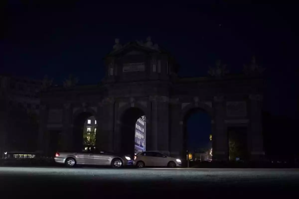 Dos coches circulan frente a un monumento histórico iluminado tenuemente por la noche.
