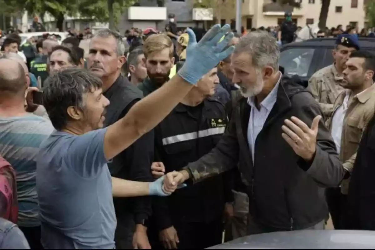 Un grupo de personas se encuentra reunido al aire libre, donde un hombre con guantes azules parece estar dialogando con otro hombre de cabello canoso y barba, mientras varias personas observan la escena.