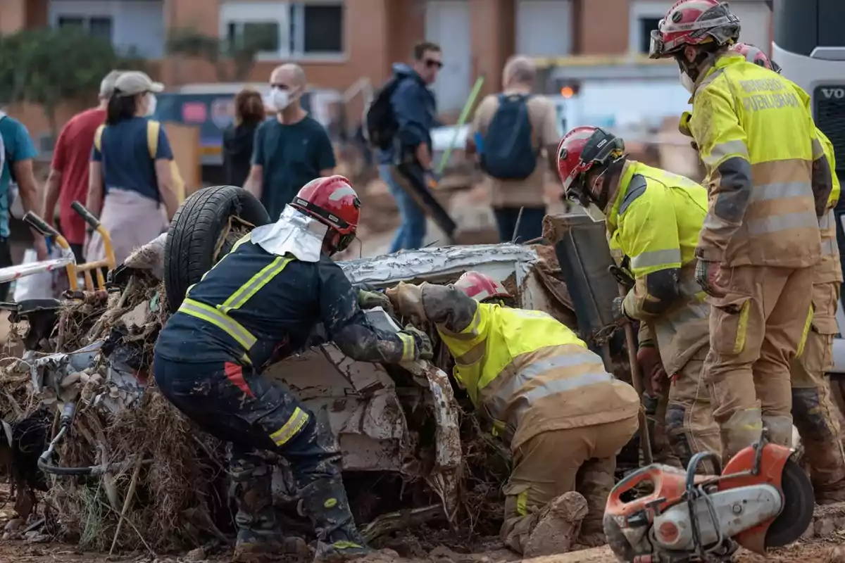 Un grupo de bomberos trabaja en el rescate de un vehículo volcado y cubierto de escombros mientras varias personas observan la escena en el fondo.