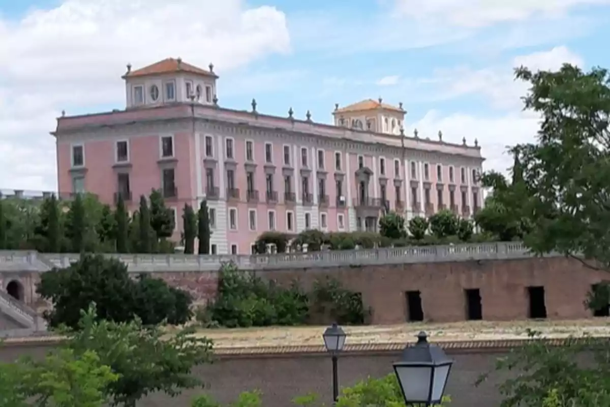 Un edificio histórico de color rosa con detalles arquitectónicos clásicos y un tejado de tejas naranjas, rodeado de vegetación y con un cielo parcialmente nublado.