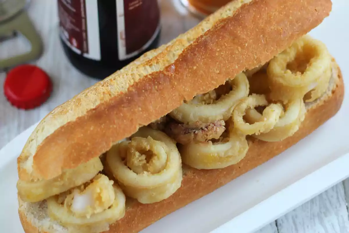 Un bocadillo de calamares fritos servido en un plato blanco con una botella y una tapa roja al fondo. Un bocadillo de calamares fritos servido en un plato blanco con una botella y una tapa roja al fondo.