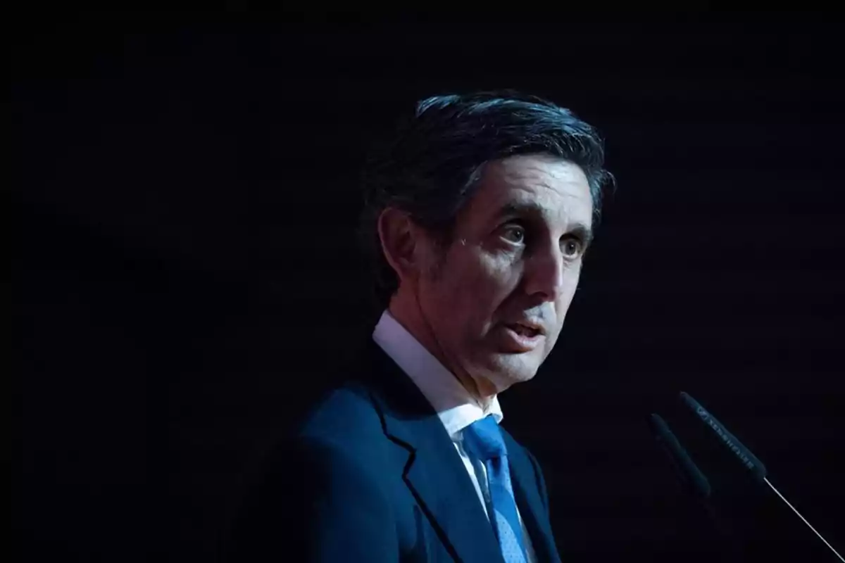Un hombre con traje oscuro y corbata azul hablando frente a un micrófono en un entorno oscuro. Un hombre con traje oscuro y corbata azul hablando frente a un micrófono en un entorno oscuro.
