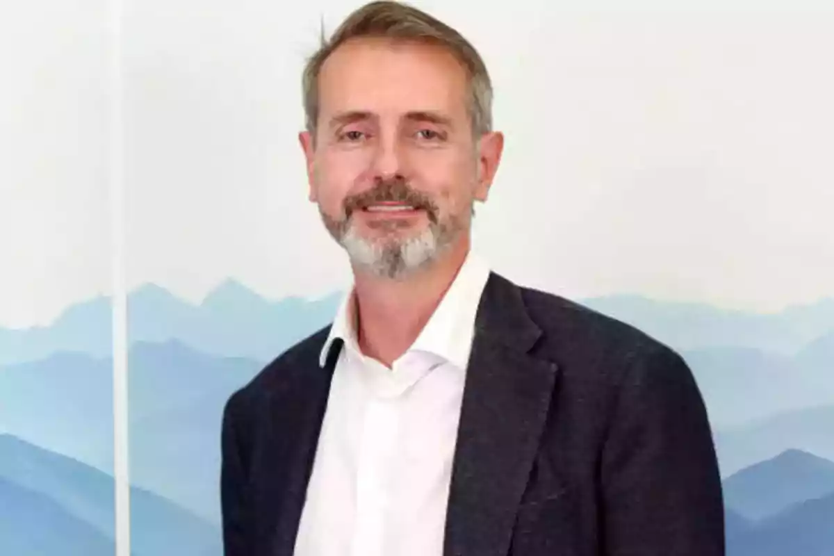 Un hombre con barba y cabello corto viste una chaqueta oscura y camisa blanca, posando frente a un fondo de montañas azules.