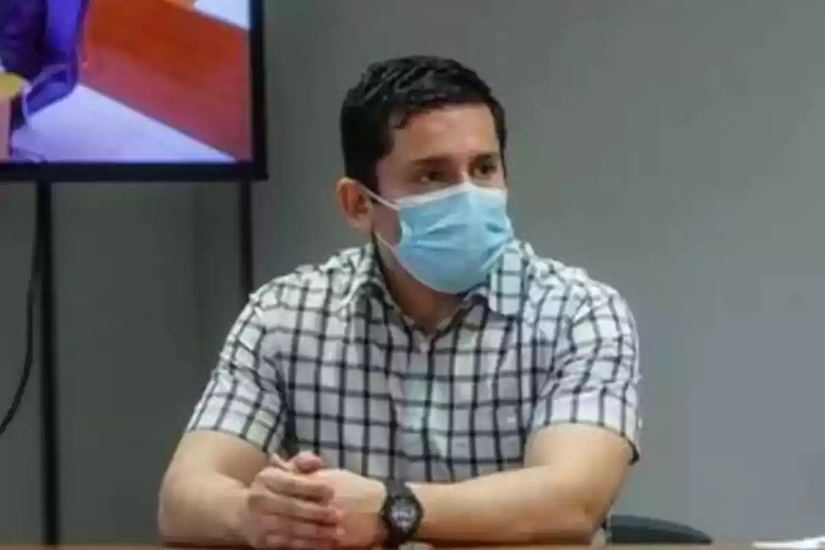 Hombre con camisa a cuadros y mascarilla sentado en una mesa.