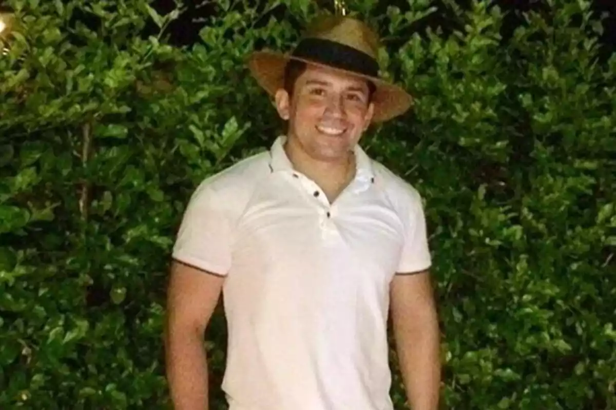 Hombre con sombrero y camisa blanca sonriendo frente a un seto verde.