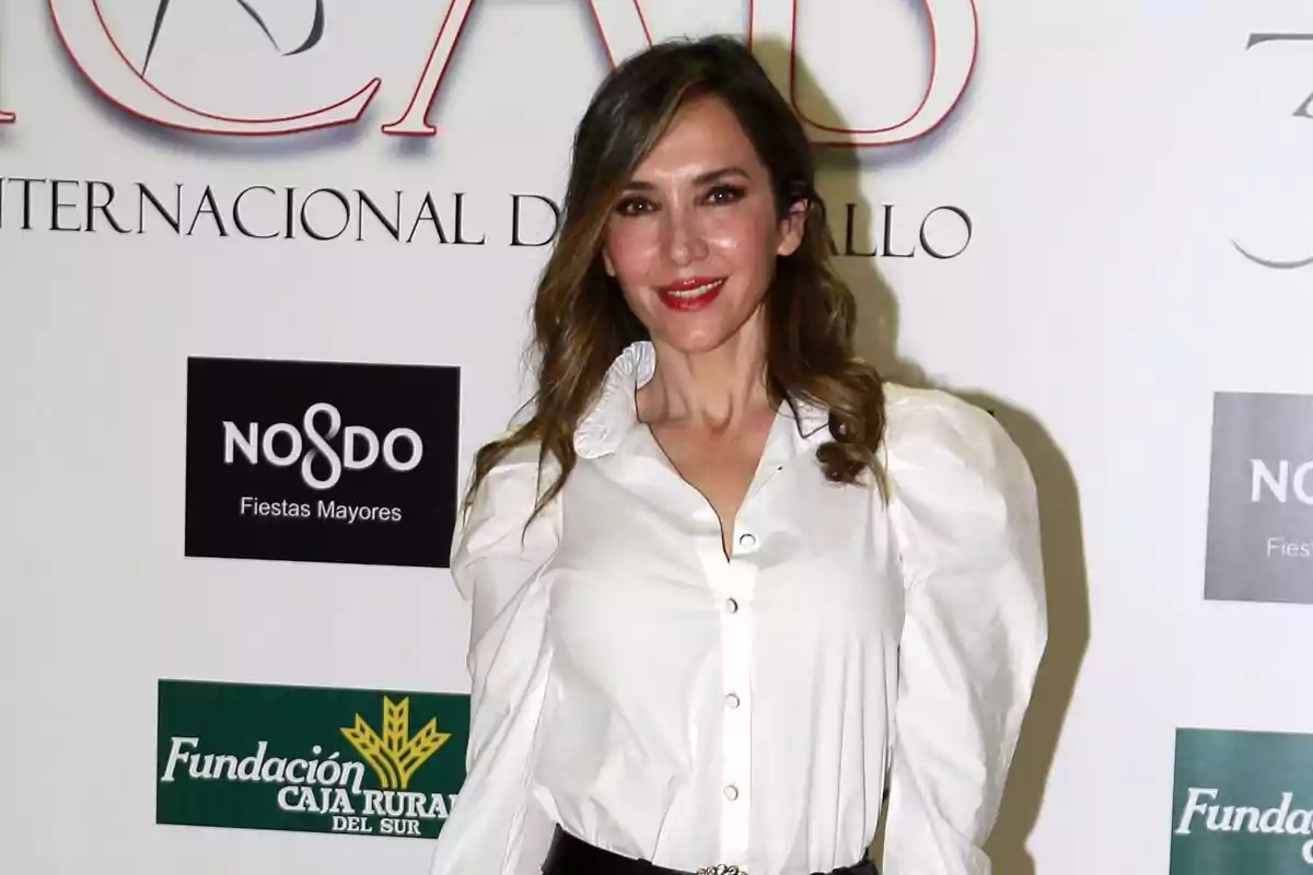 Mujer con cabello suelto y ondulado vestida con blusa blanca posando frente a un fondo con logotipos y texto Mujer con cabello suelto y ondulado vestida con blusa blanca posando frente a un fondo con logotipos y texto