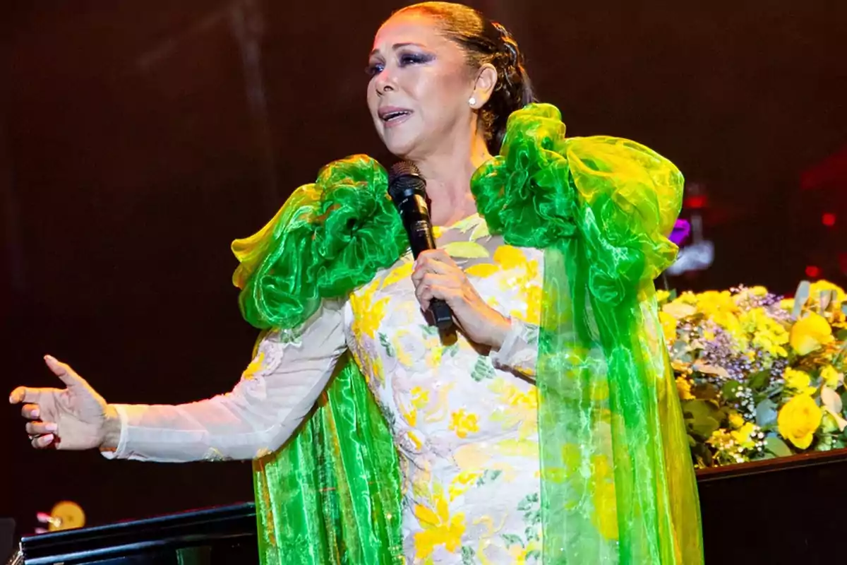 Una mujer con un vestido colorido y una capa verde canta en un escenario mientras sostiene un micrófono.