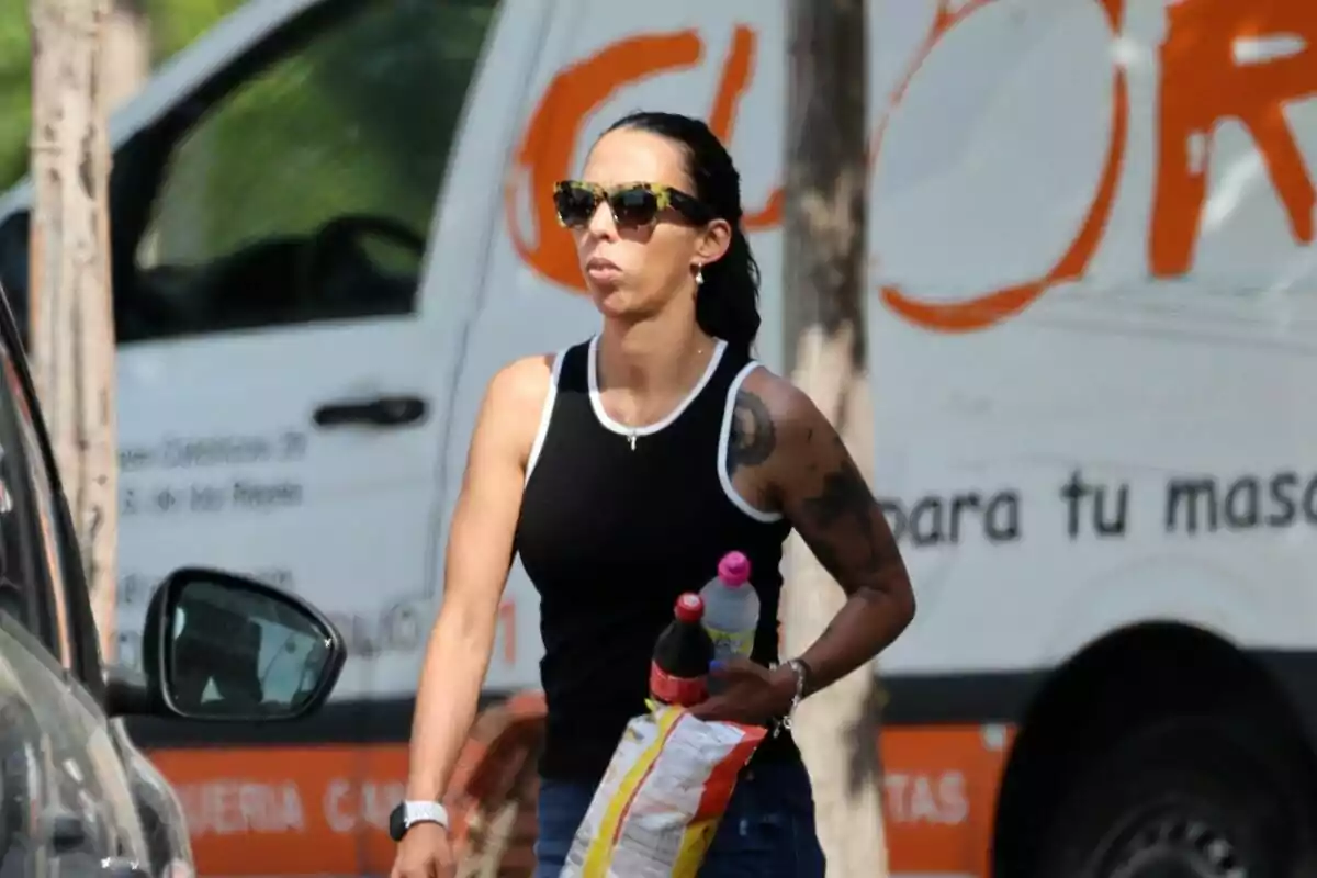 Paola Olmedo seria con el pelo recogido, gafas de sol, tirantes negros y blancos y dos botellas y una bolsa de patatas en la mano Paola Olmedo seria con el pelo recogido, gafas de sol, tirantes negros y blancos y dos botellas y una bolsa de patatas en la mano