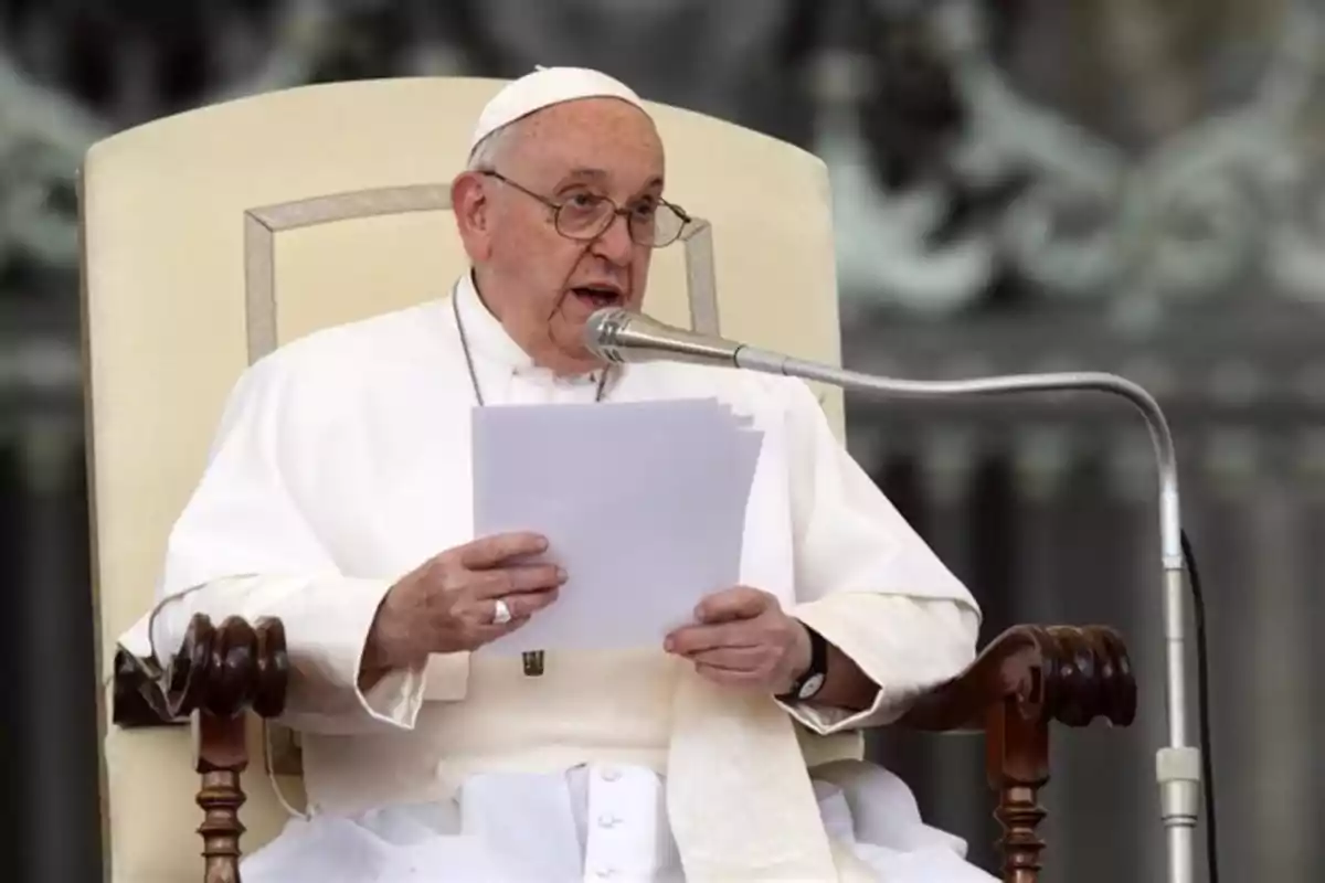 Un hombre vestido con atuendo religioso blanco está sentado en una silla, sosteniendo papeles y hablando frente a un micrófono.