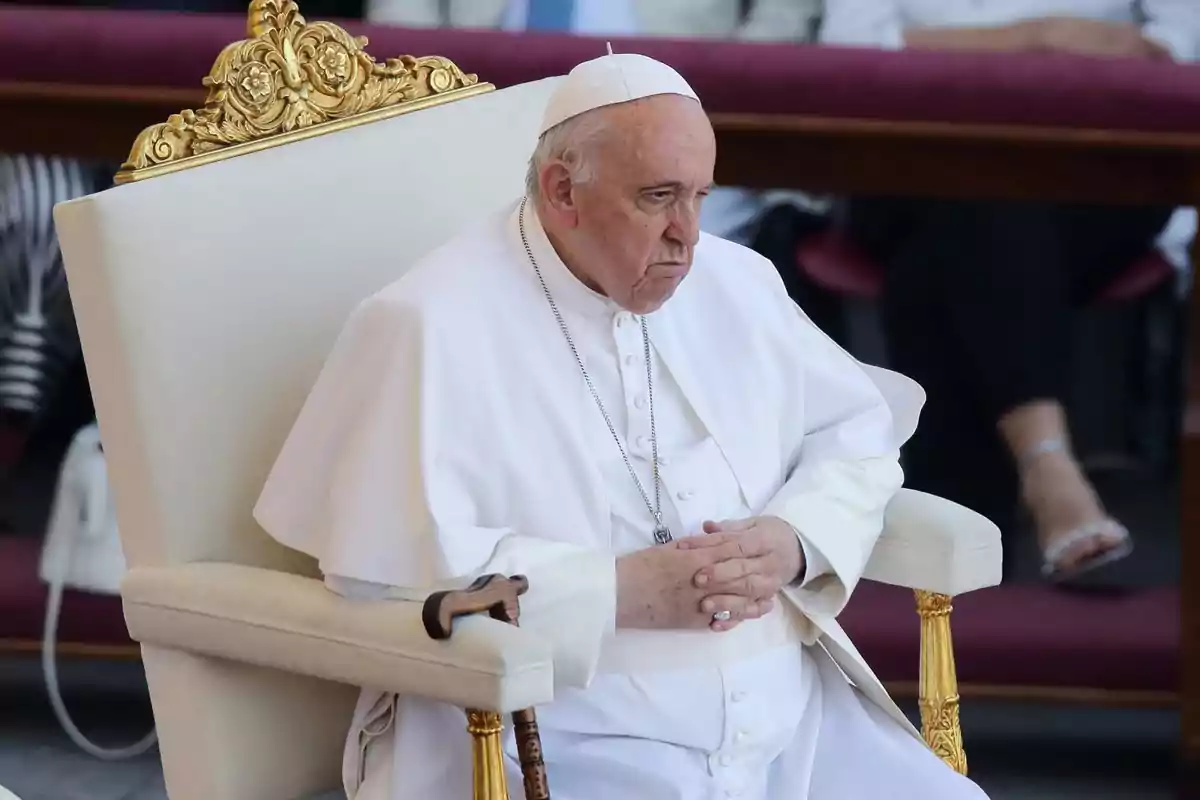 Un hombre mayor vestido con atuendo religioso blanco está sentado en una silla con detalles dorados, con las manos entrelazadas y un bastón apoyado en el brazo de la silla.