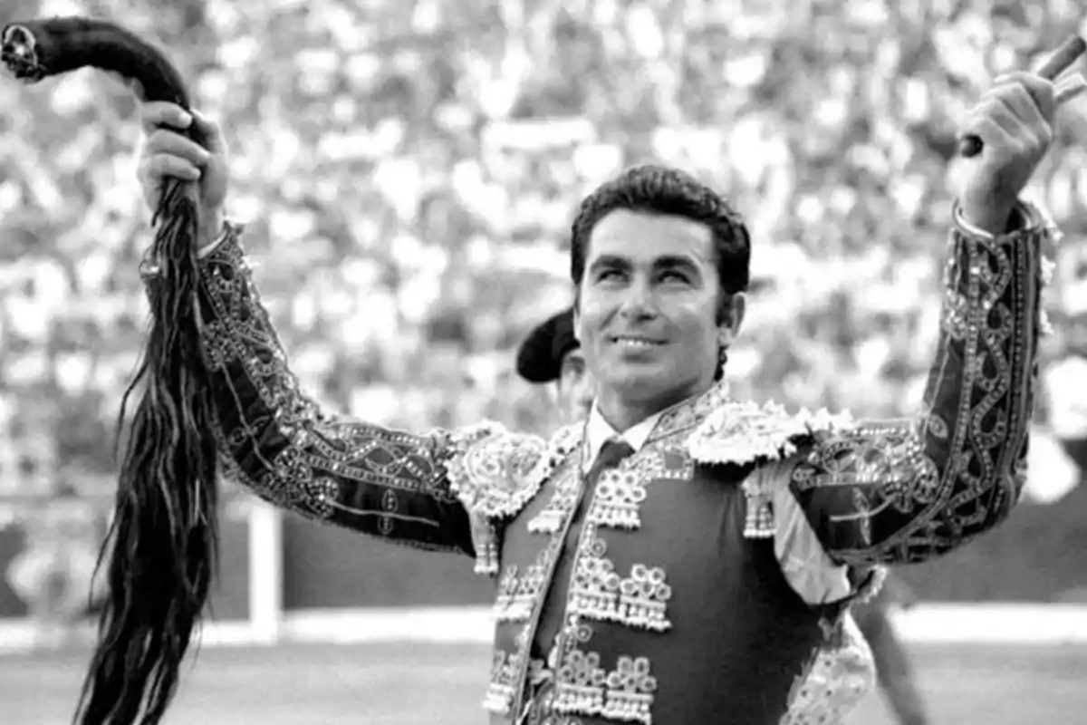 Un torero sonriente levanta un trofeo en una plaza de toros llena de espectadores.