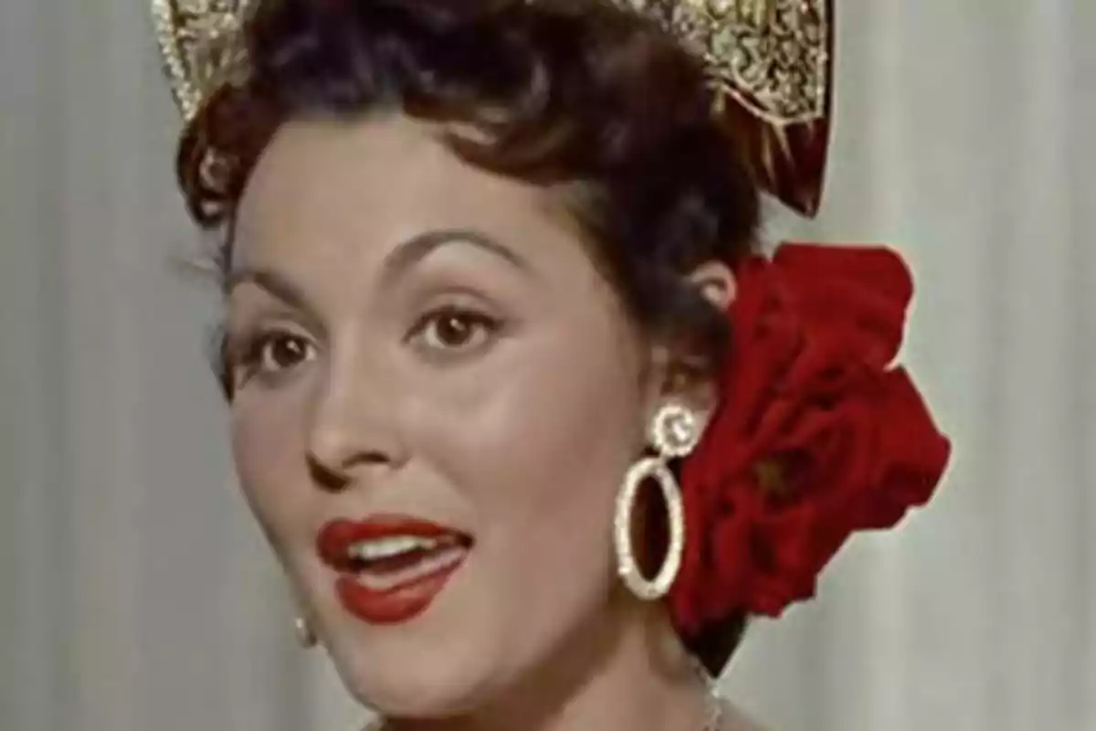 Una mujer con un tocado dorado y una flor roja en el cabello, usando aretes grandes y redondos, con los labios pintados de rojo.