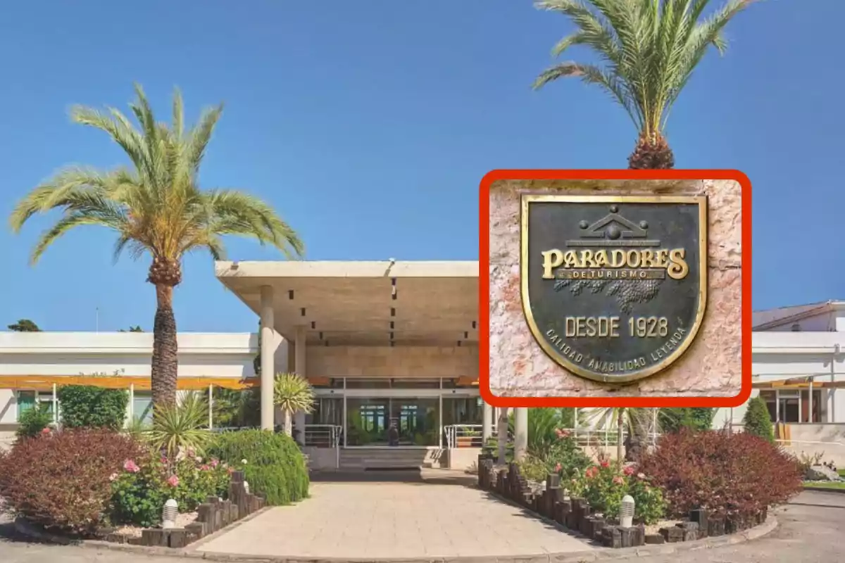 Entrada de un edificio con palmeras y un jardín bien cuidado, con un cartel que dice "Paradores de Turismo, desde 1928, calidad, amabilidad, leyenda".