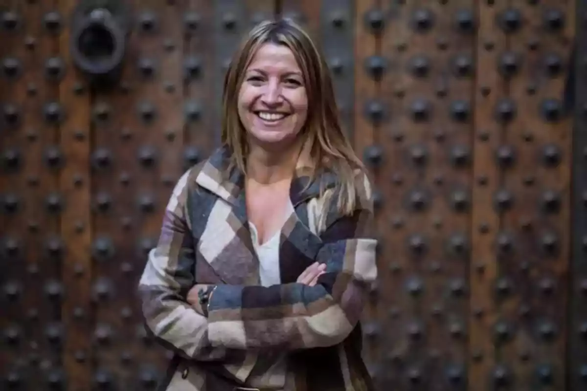 Una mujer sonriente con los brazos cruzados frente a una puerta de madera con detalles metálicos.