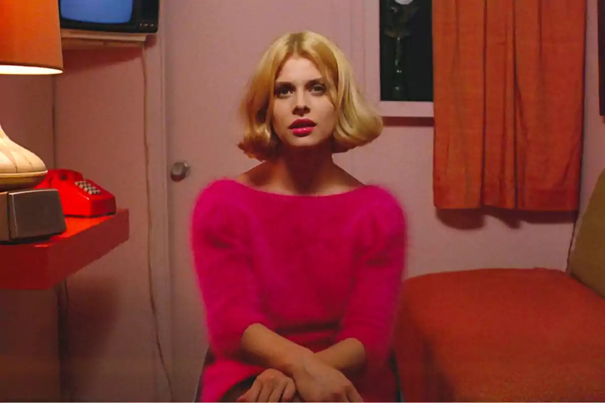 Una persona con cabello rubio y un suéter rosa está sentada en una habitación con una decoración retro que incluye una lámpara, un teléfono rojo y cortinas naranjas. Una persona con cabello rubio y un suéter rosa está sentada en una habitación con una decoración retro que incluye una lámpara, un teléfono rojo y cortinas naranjas.