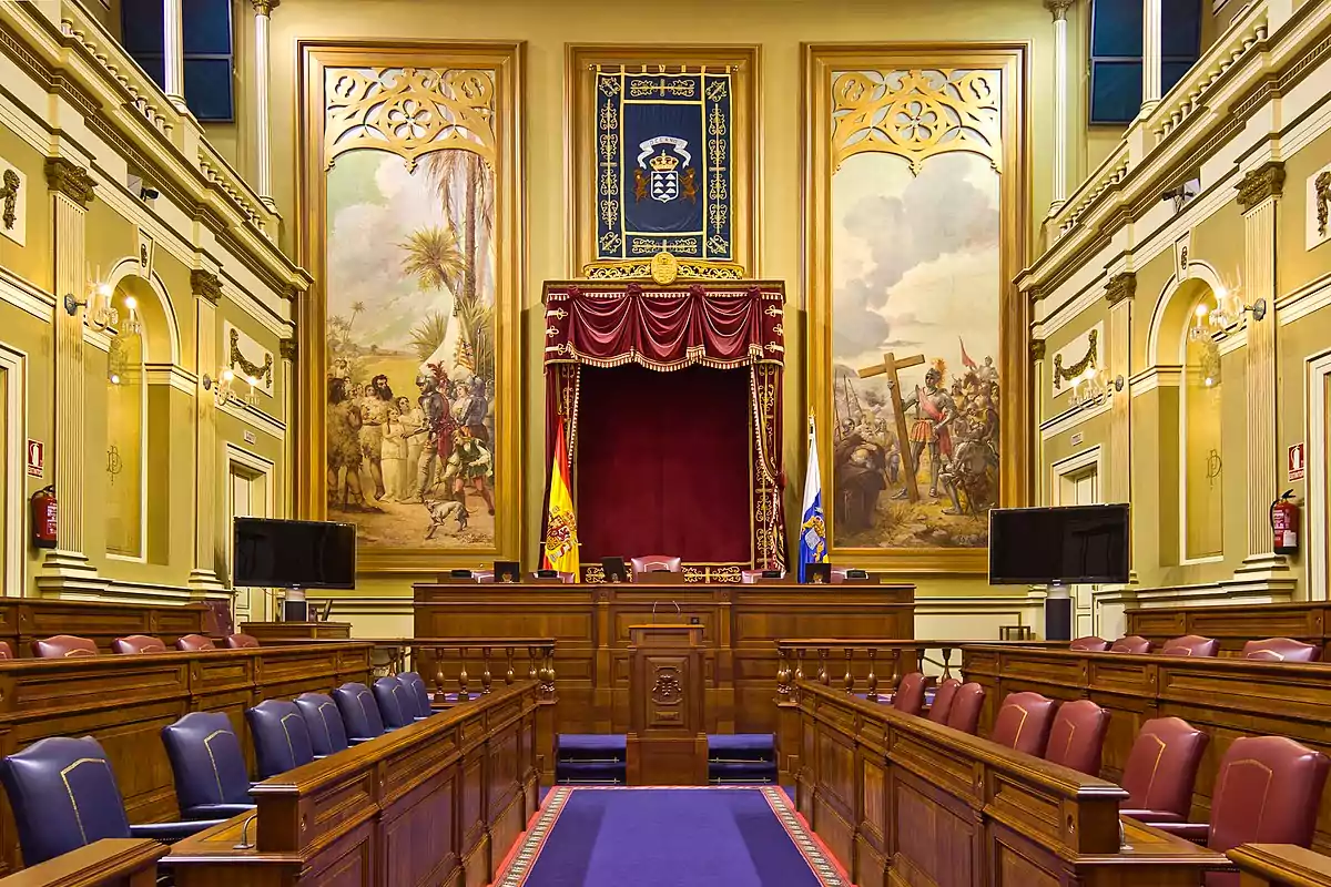Sala de un parlamento con asientos de madera y cuero, un estrado central con cortinas rojas, banderas y grandes murales en las paredes.