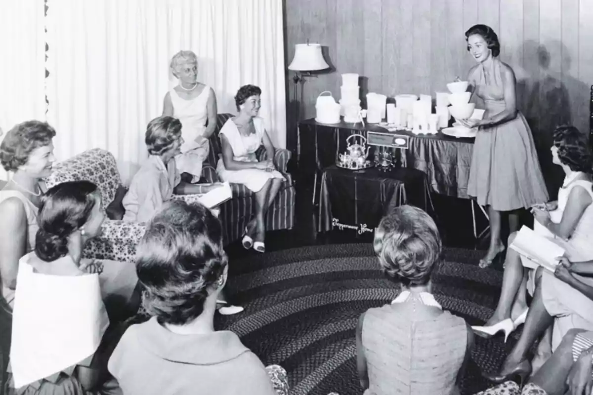 Un grupo de mujeres se encuentra reunido en una sala, mientras una de ellas, de pie, les muestra varios recipientes de plástico sobre una mesa.