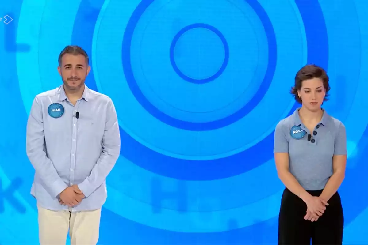Juan y Rosa de pie en un plató de televisión con fondo azul y círculos concéntricos, ambos llevan chapas con sus nombres y miran al frente. Captura de 'Pasapalabra' Juan y Rosa de pie en un plató de televisión con fondo azul y círculos concéntricos, ambos llevan chapas con sus nombres y miran al frente. Captura de 'Pasapalabra'