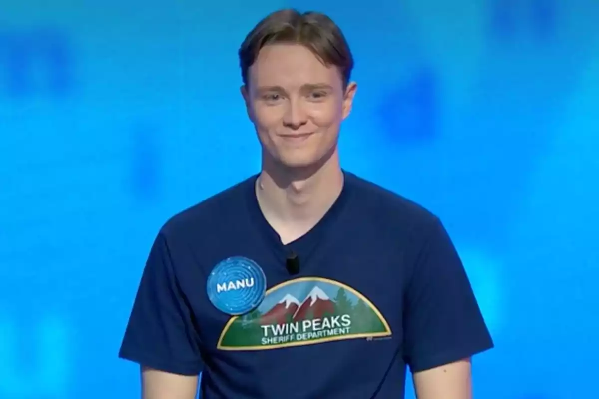 Manu con una camiseta azul que tiene un diseño de Twin Peaks Sheriff Department y una etiqueta que dice Manu sonríe frente a un fondo azul. Captura de 'Pasapalabra' Manu con una camiseta azul que tiene un diseño de Twin Peaks Sheriff Department y una etiqueta que dice Manu sonríe frente a un fondo azul. Captura de 'Pasapalabra'