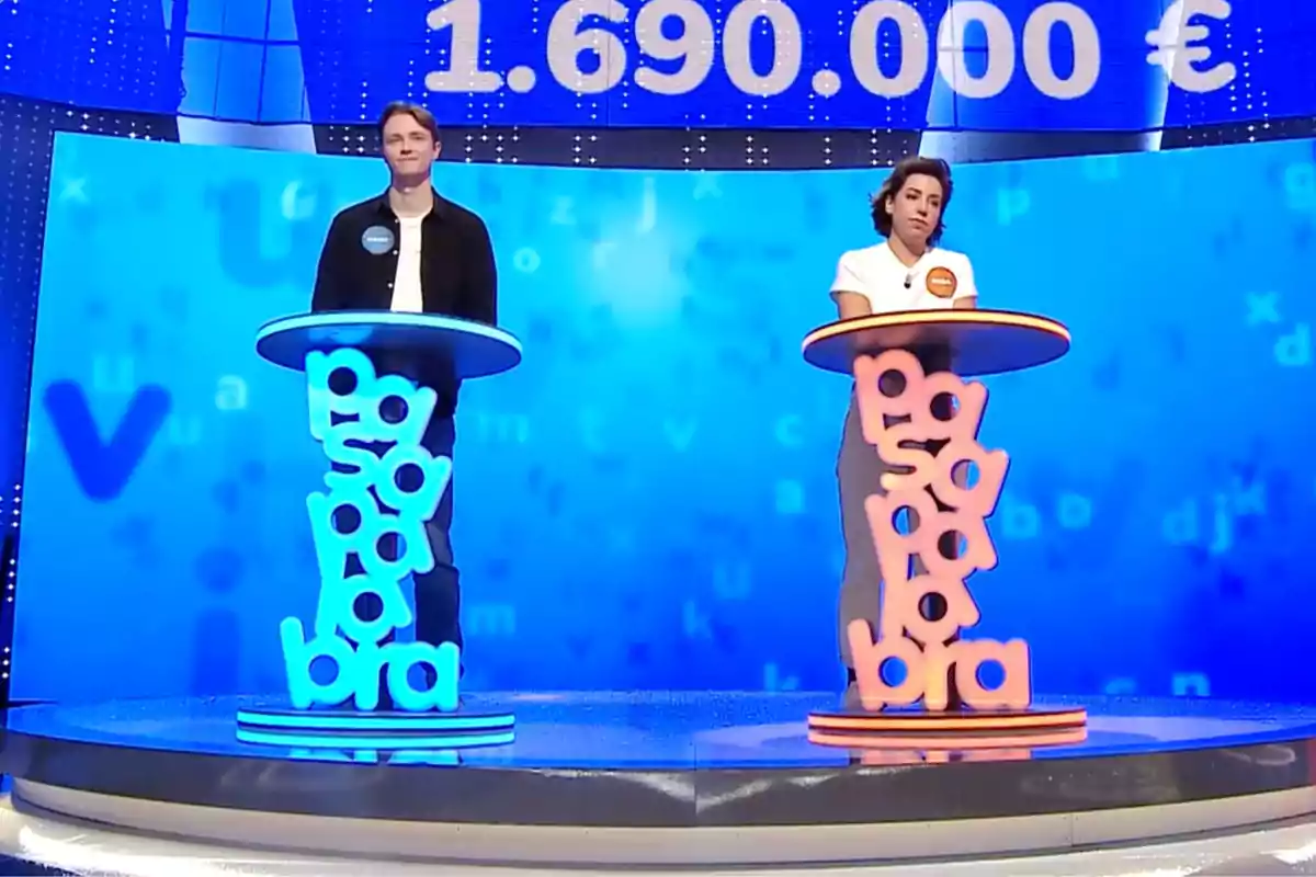 Manu y Rosa están de pie en sus respectivos atriles, uno azul y otro naranja, en un plató de televisión con un fondo azul y una cifra grande de dinero en la parte superior. Captura de 'Pasapalabra' Manu y Rosa están de pie en sus respectivos atriles, uno azul y otro naranja, en un plató de televisión con un fondo azul y una cifra grande de dinero en la parte superior. Captura de 'Pasapalabra'