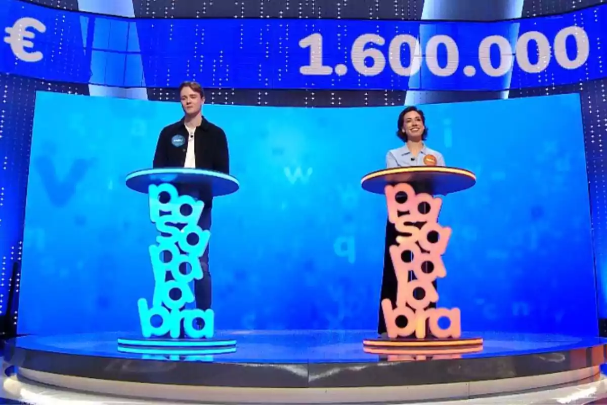 Manu y Rosa están de pie en un plató de televisión con un fondo azul, cada uno detrás de un podio que tiene la palabra Manu y Rosa están de pie en un plató de televisión con un fondo azul, cada uno detrás de un podio que tiene la palabra