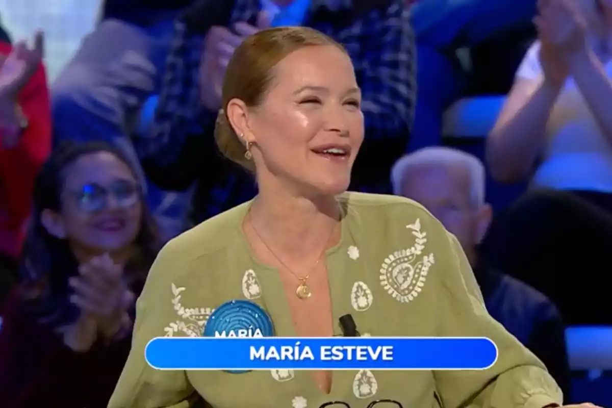 María Esteve sonriente con cabello recogido y blusa verde claro participando en un programa de televisión con público aplaudiendo al fondo y un letrero azul con el nombre María Esteve. Captura de 'Pasapalabra' María Esteve sonriente con cabello recogido y blusa verde claro participando en un programa de televisión con público aplaudiendo al fondo y un letrero azul con el nombre María Esteve. Captura de 'Pasapalabra'