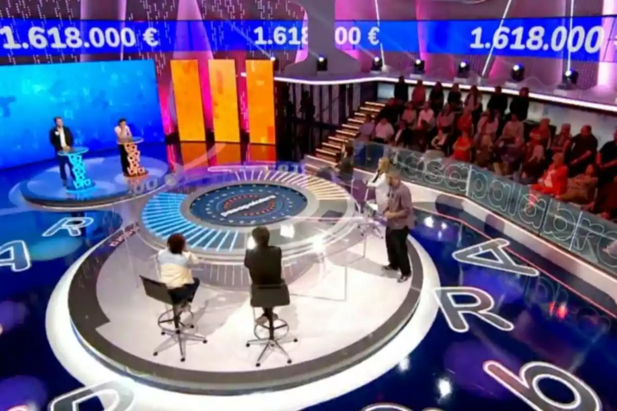Un plató de concurso televisivo con un diseño colorido y moderno, donde se observan participantes en sus respectivos podios, un presentador y una audiencia sentada al fondo, con una pantalla que muestra una cantidad de dinero. Captura de 'Pasapalabra' Un plató de concurso televisivo con un diseño colorido y moderno, donde se observan participantes en sus respectivos podios, un presentador y una audiencia sentada al fondo, con una pantalla que muestra una cantidad de dinero. Captura de 'Pasapalabra'