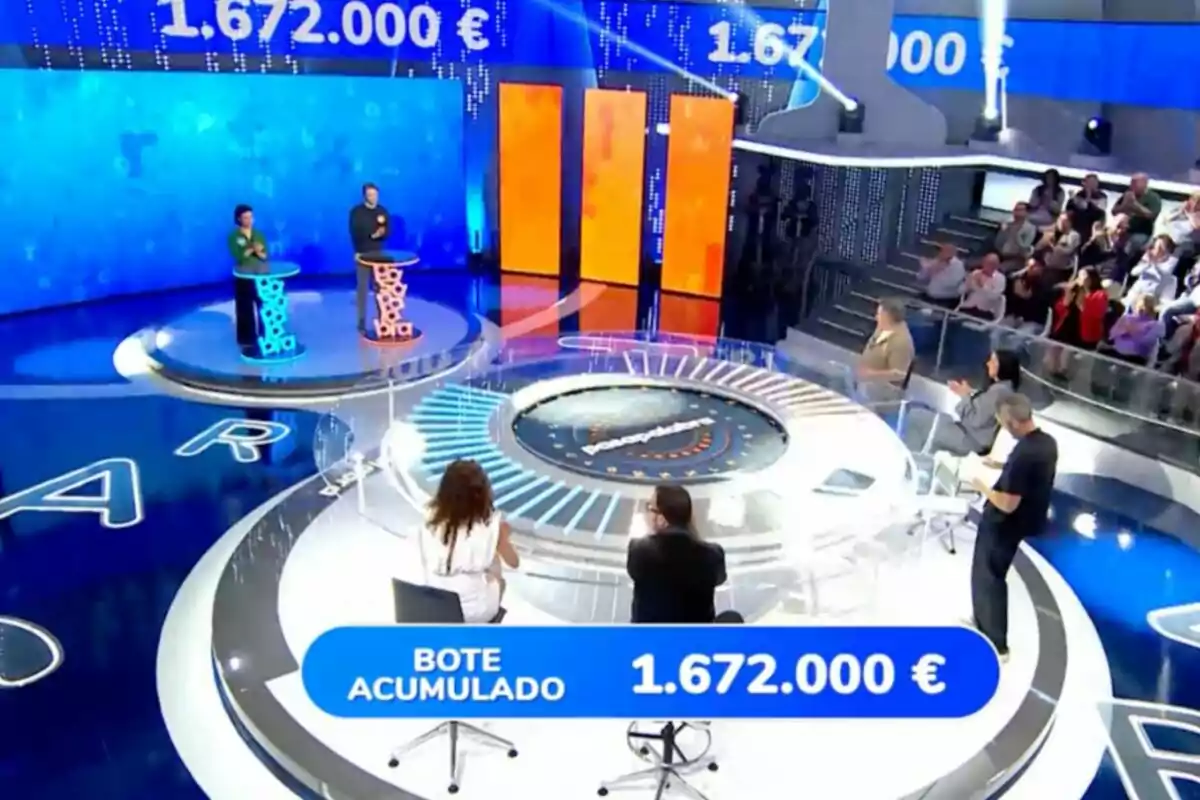 Escenario de un concurso televisivo con dos concursantes de pie en atriles iluminados, un panel con cifras grandes en euros y público observando desde las gradas. Captura de 'Pasapalabra' Escenario de un concurso televisivo con dos concursantes de pie en atriles iluminados, un panel con cifras grandes en euros y público observando desde las gradas. Captura de 'Pasapalabra'