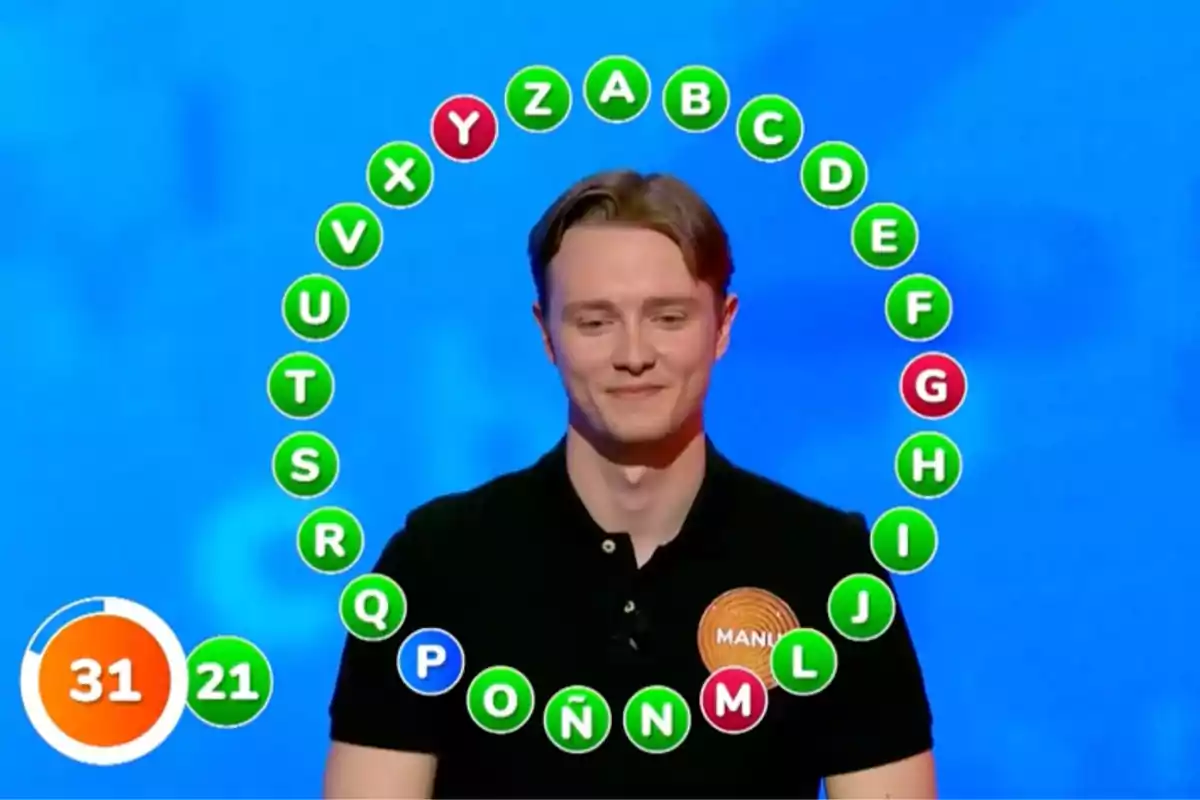 Manu en un programa de televisión está de pie frente a una pantalla azul con letras del alfabeto en círculos de colores a su alrededor y un marcador de tiempo en la esquina inferior izquierda. Captura de 'Pasapalabra' Manu en un programa de televisión está de pie frente a una pantalla azul con letras del alfabeto en círculos de colores a su alrededor y un marcador de tiempo en la esquina inferior izquierda. Captura de 'Pasapalabra'