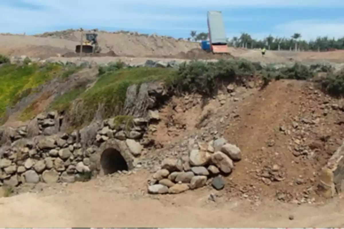 Túnel de piedra parcialmente cubierto por tierra y vegetación con maquinaria de construcción trabajando en el fondo Túnel de piedra parcialmente cubierto por tierra y vegetación con maquinaria de construcción trabajando en el fondo