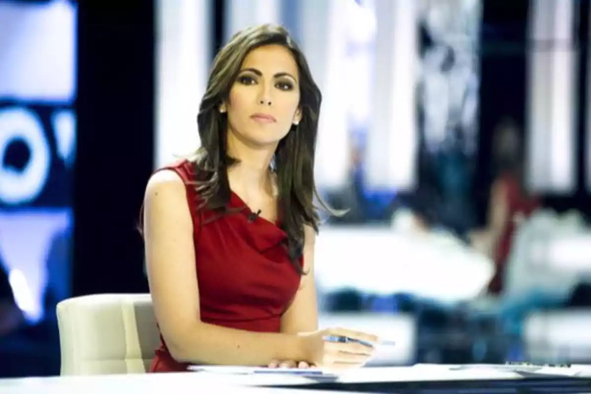Woman in a red dress sitting at a television studio desk with a serious expression and a pen in her hand Woman in a red dress sitting at a television studio desk with a serious expression and a pen in her hand