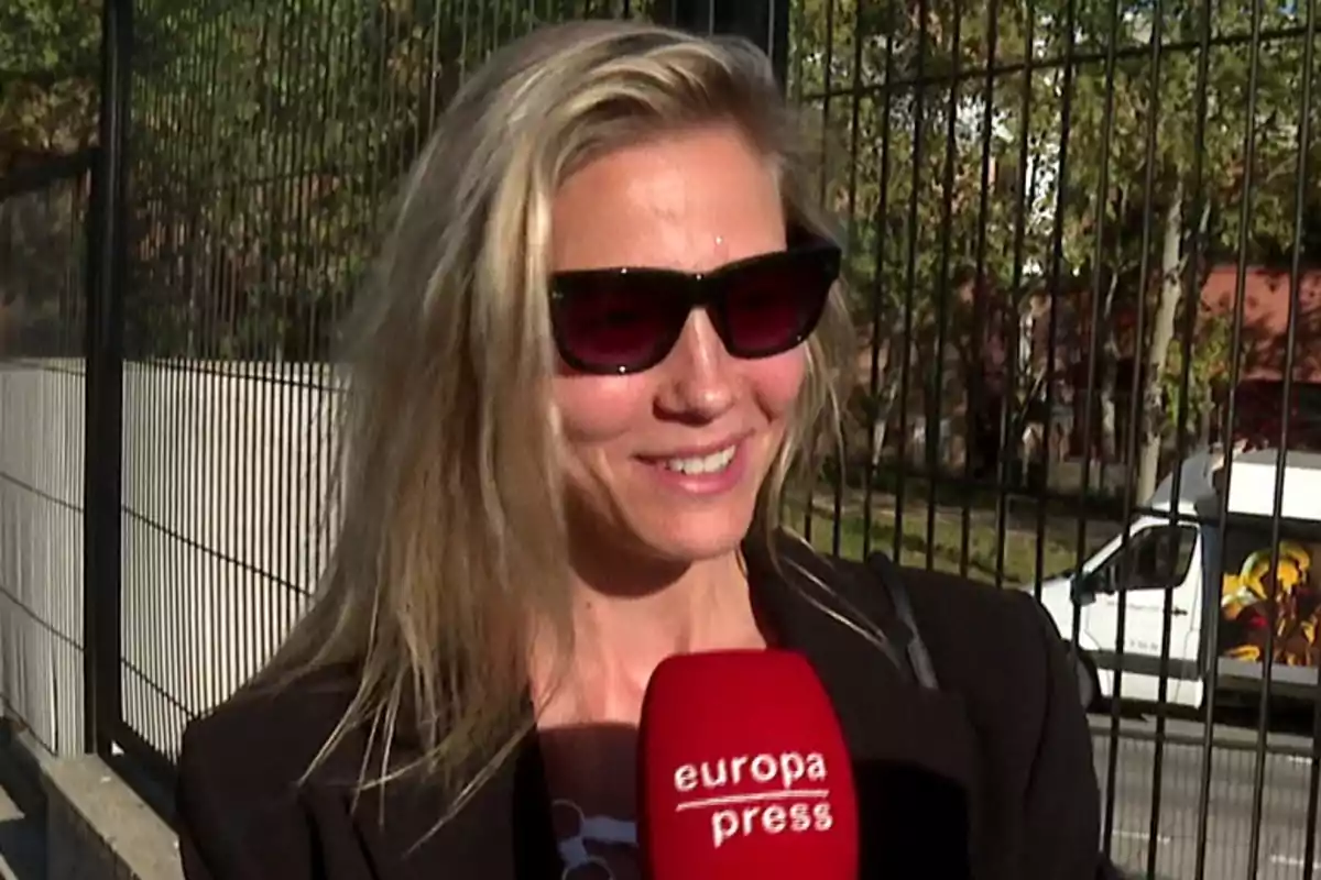 Patricia Montero con gafas de sol sonríe mientras sostiene un micrófono de Europa Press frente a una reja y un vehículo estacionado. Patricia Montero con gafas de sol sonríe mientras sostiene un micrófono de Europa Press frente a una reja y un vehículo estacionado.