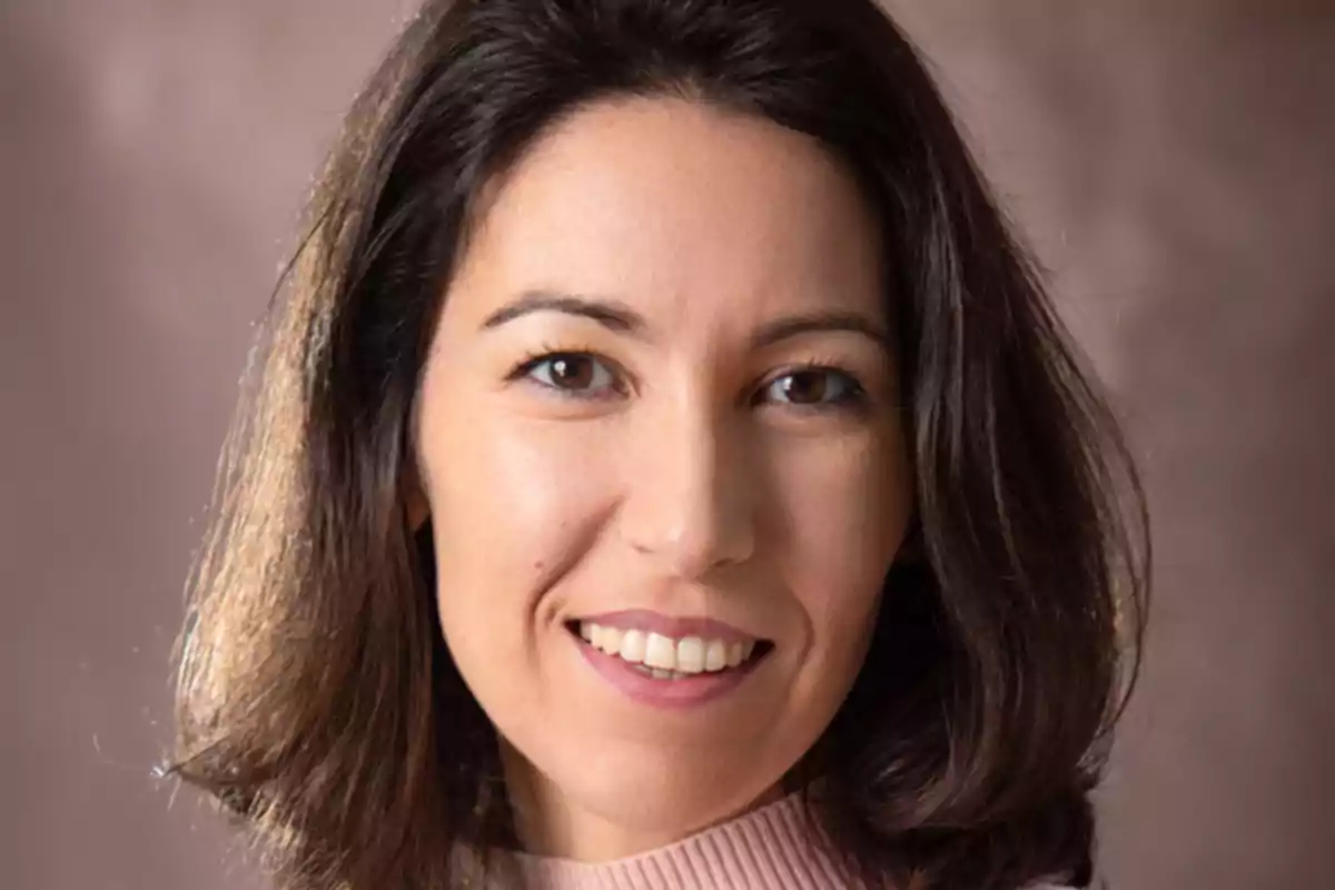 Retrato de una mujer con cabello oscuro y lacio, sonriendo y mirando a la cámara, con un fondo difuminado.