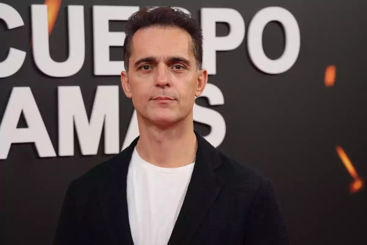 Un hombre con cabello oscuro y corto lleva una camiseta blanca y una chaqueta oscura posando frente a un fondo con letras grandes.