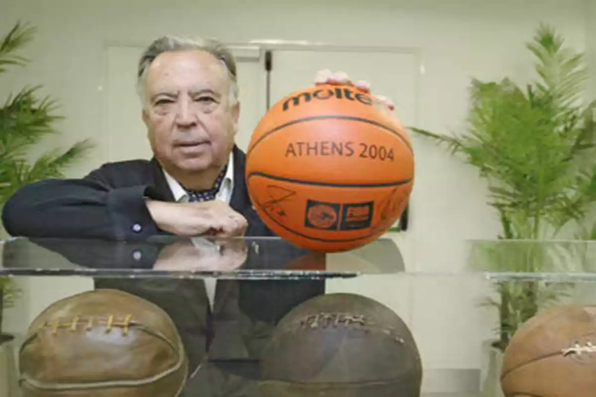 Un hombre mayor sostiene un balón de baloncesto de los Juegos Olímpicos de Atenas 2004, mientras está de pie detrás de una vitrina que contiene balones de baloncesto antiguos.