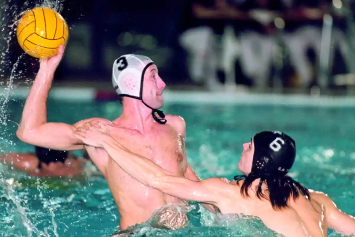 Dos jugadores de waterpolo compiten en una piscina, uno de ellos sostiene el balón mientras el otro intenta bloquearlo.