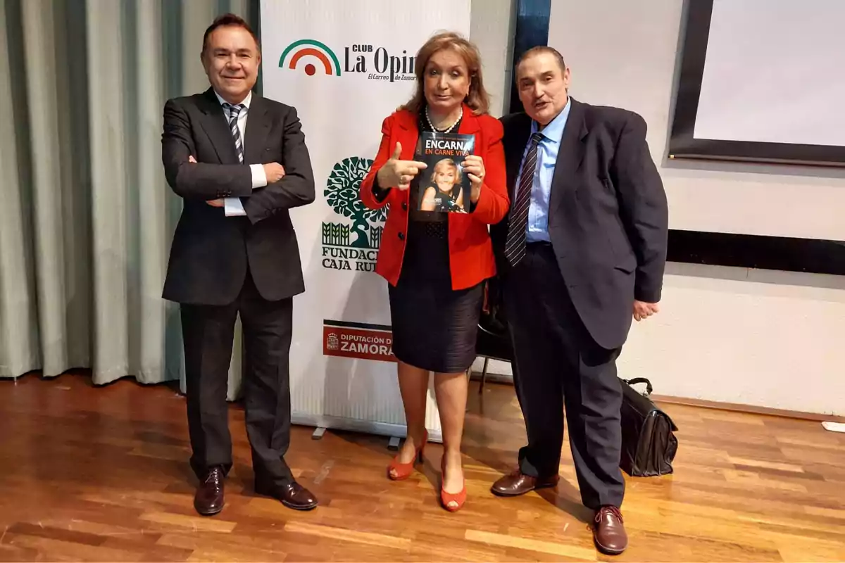 Tres personas posan para una foto, una mujer en el centro sostiene un libro titulado 