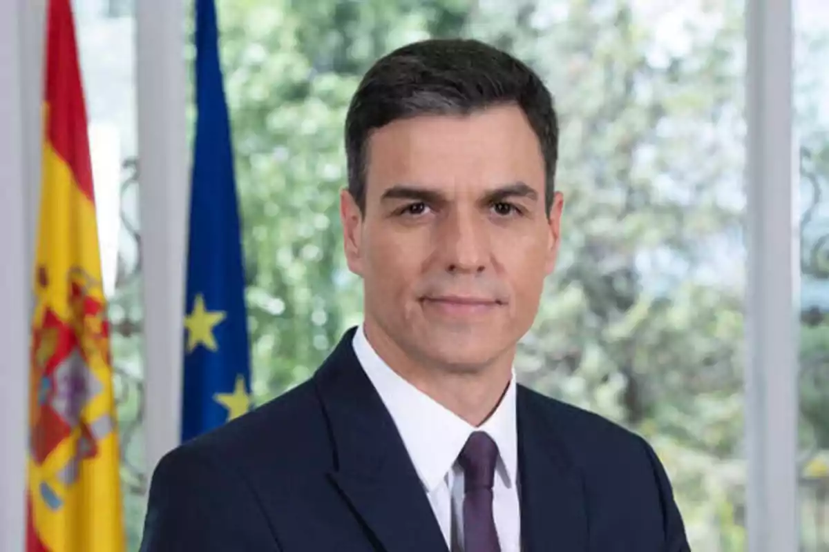 Hombre con traje oscuro y corbata, de pie frente a las banderas de España y la Unión Europea.