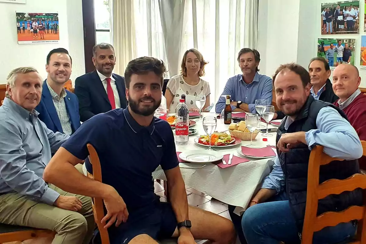 Un grupo de personas sentadas alrededor de una mesa redonda con comida y bebidas, en un ambiente interior con cuadros en la pared.