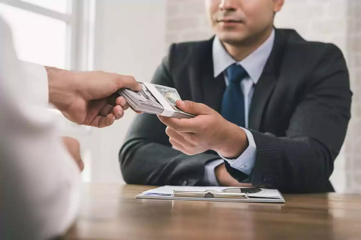 Un hombre en traje recibe un fajo de billetes de otra persona sobre una mesa con un portapapeles.