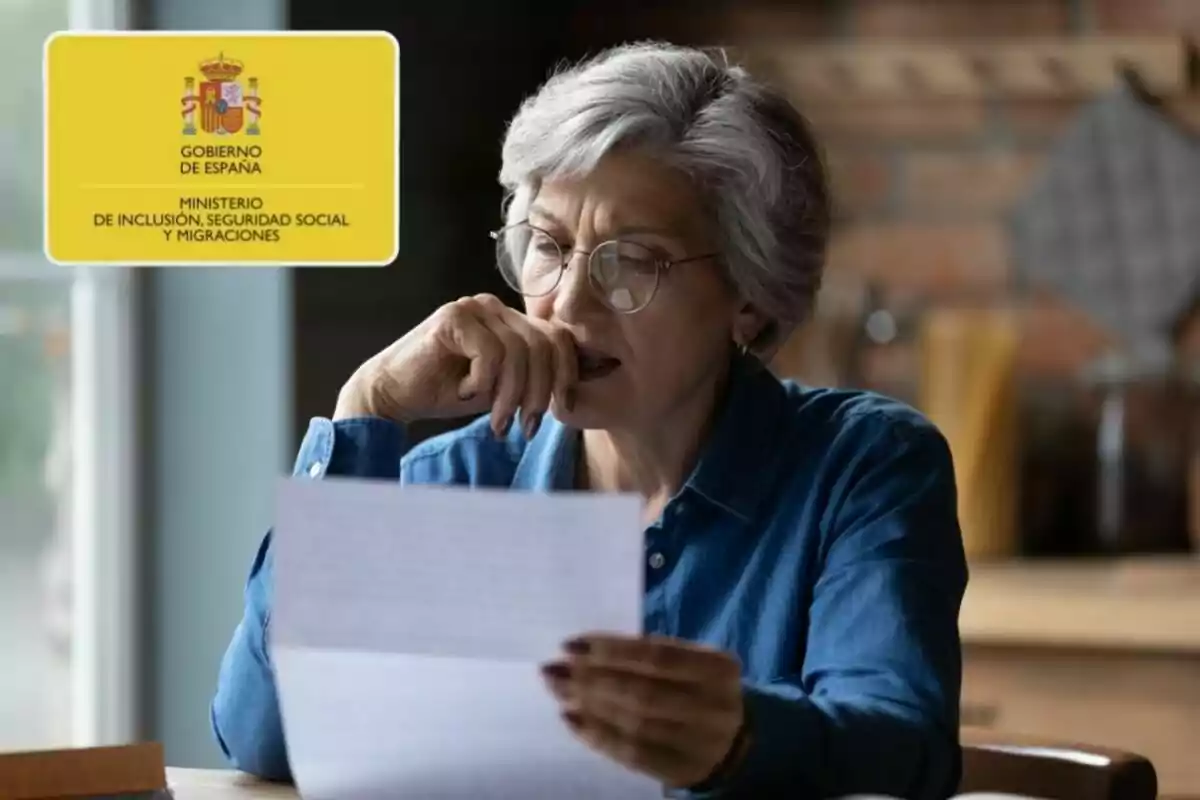 Una mujer mayor con gafas lee un documento en una mesa, con un cartel del Gobierno de España y el Ministerio de Inclusión, Seguridad Social y Migraciones en la esquina superior izquierda.