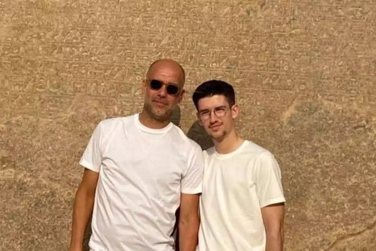 Dos personas con camisetas blancas posan frente a una pared con jeroglíficos.