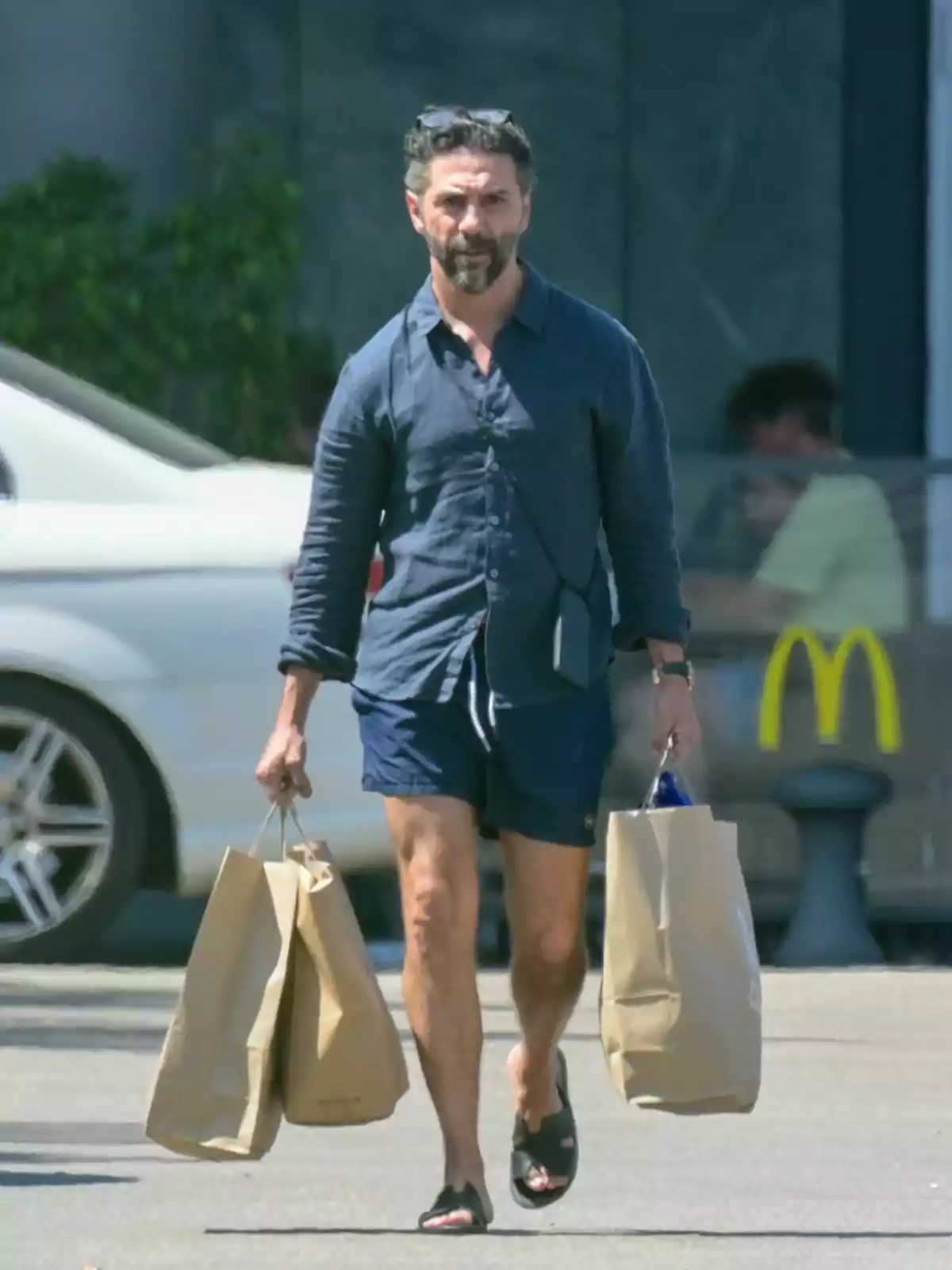 Pepe Bastón con camisa azul y pantalones cortos lleva dos bolsas de papel mientras camina por un estacionamiento.