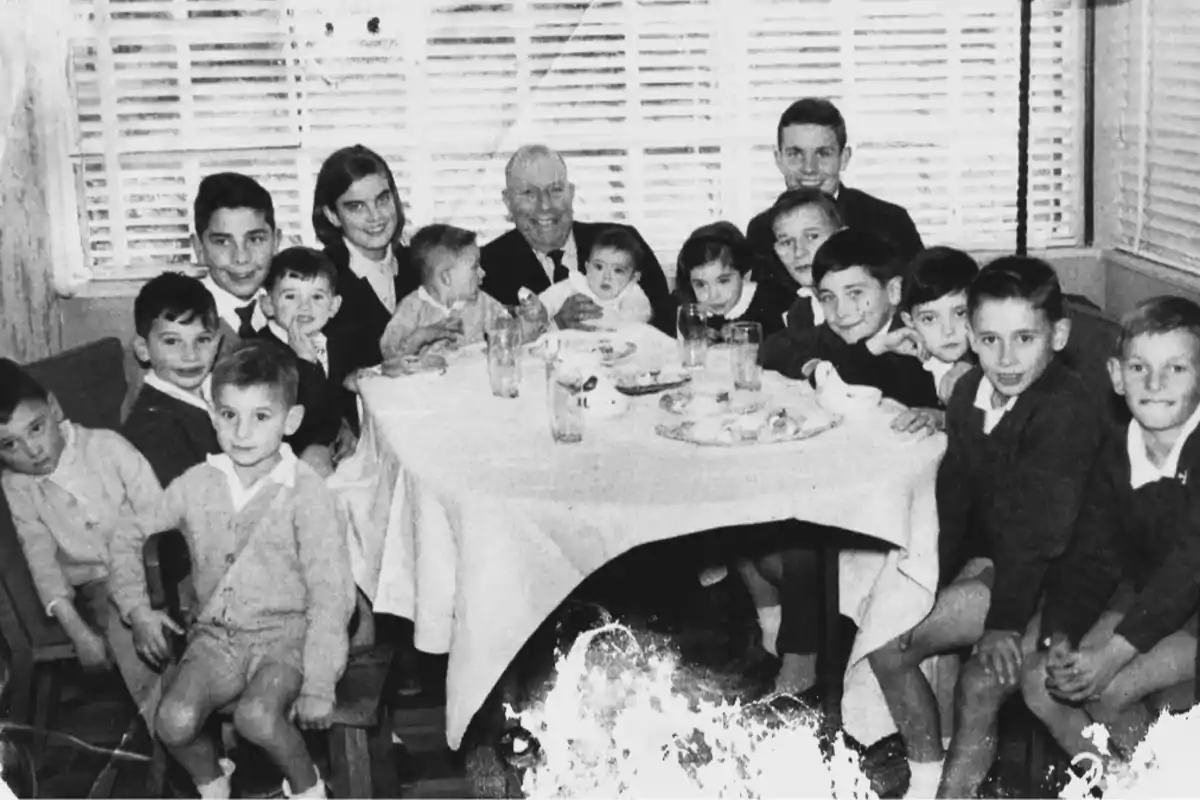 Un grupo de personas, incluyendo niños y un adulto mayor, están reunidos alrededor de una mesa en un ambiente interior.