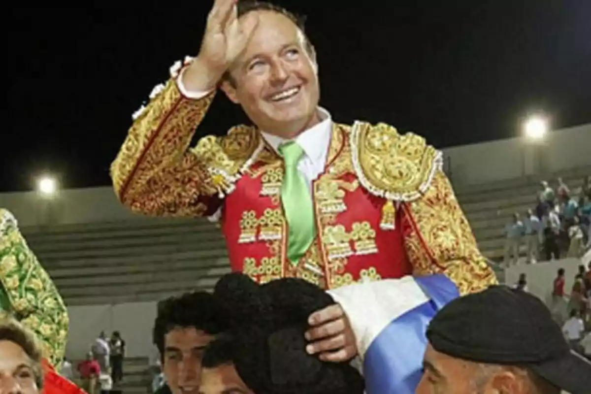 Pepe Luis Vázquez Silva vestido con traje de luces rojo y dorado saluda mientras es llevado en hombros por varias personas en una plaza de toros.