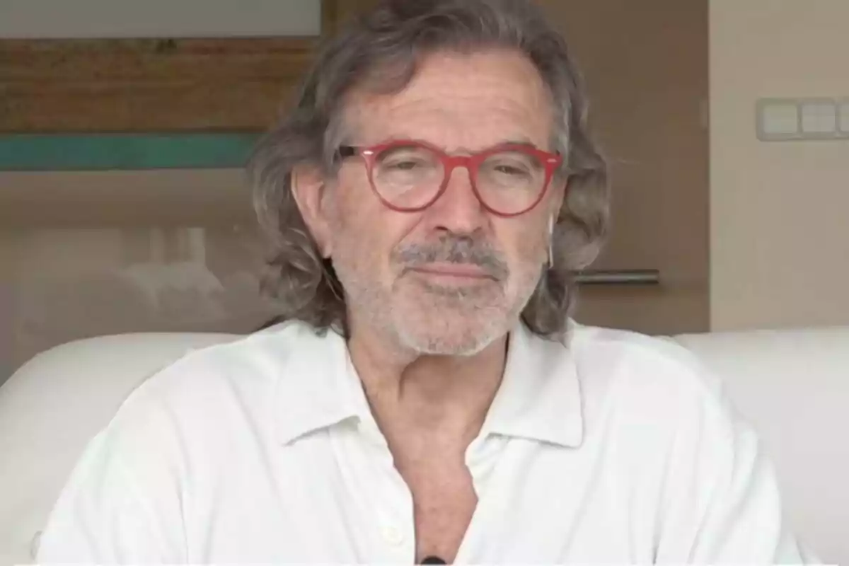 Plano corto de Pepe Navarro con gafas rojas y cabello canoso lleva una camisa blanca mientras está sentado en un sofá.